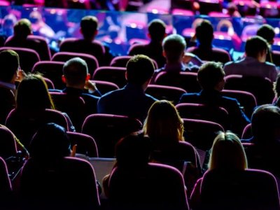 Backs of an audience in a dark conference room facing the stage.